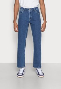 Blauwe denim jeans met een rechte pijp, voorzien van een knoopsluiting, vijf zakken en subtiele vervaging. Draagt wit- en paarskleurige sneakers.