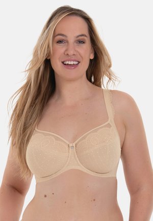 Glimlachende vrouw met lang blond haar, gekleed in een beige kanten en mesh ondersteunende beha, tegen een effen lichte achtergrond.