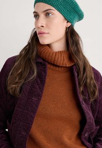 Fioletowa kurtka z korduroyu z pikowanym wykończeniem na pomarańczowym swetrze z golfem, zestawiona z zieloną wełnianą czapką. Gładkie faktury i kontrastujące kolory.