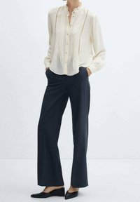 Crèmekleurige blouse met een gerimpelde kraag en verticale plooien, gecombineerd met donkergekleurde wijde broeken en zwarte instapschoenen. Soepele textuur, neutrale kleur.