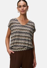 Kvinde iført en kortærmet bluse med zigzag-mønster og V-udskæring samt brune bukser, stående med den ene hånd i lommen.