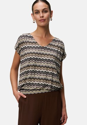 Donna che indossa una blusa a maniche corte con motivo a zigzag e scollatura a V, con pantaloni marroni, in piedi con una mano in tasca.