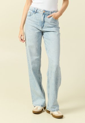 Vaqueros rectos de tiro alto en azul claro, combinados con zapatillas blancas y un top sin mangas blanco, con una mano en el bolsillo.