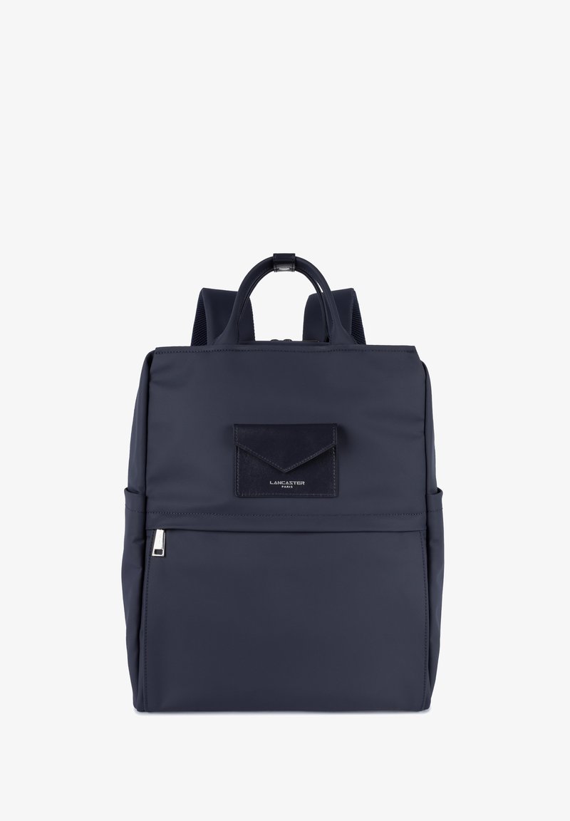 Sac à dos bleu marine avec poche zippée à l'avant, poignée supérieure, bretelles rembourrées et petite poche rectangulaire à rabat avec le logo Lancaster Paris.