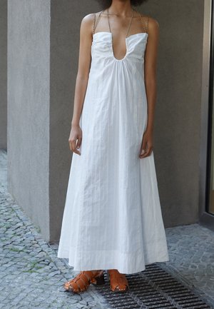 Witte mouwloze maxi-jurk met een diepe V-hals en een geplooide taille, met kettingbandjes. Gecombineerd met oranje sandalen met bandjes.