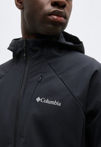 Κοντινό πλάνο ενός ατόμου που φοράει μαύρο αδιάβροχο μπουφάν Columbia με κουκούλα και φερμουάρ στην τσέπη στο στήθος.