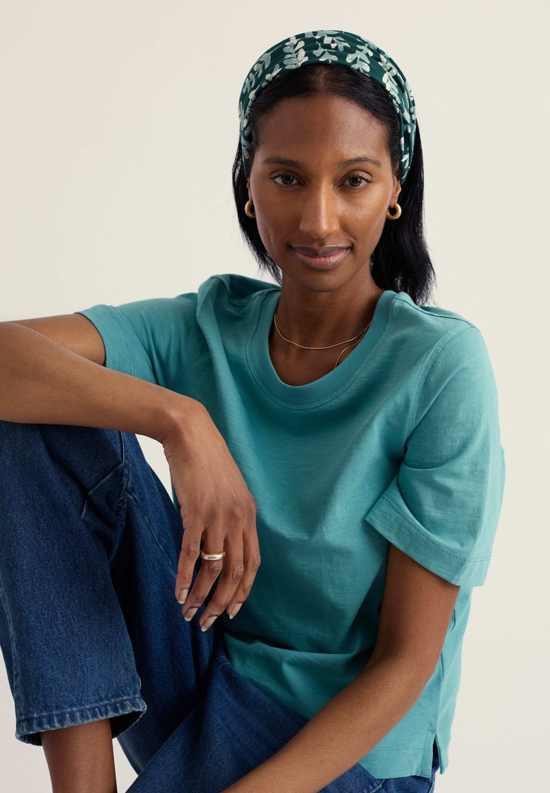 Mujer con camiseta turquesa, banda para la cabeza verde con estampado, aretes de aro dorados y jeans azules, sentada con una rodilla levantada sobre un fondo liso.