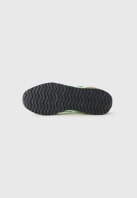 Semelle en caoutchouc noir avec un motif de semelle en zigzag et la marque "new balance" répétée sur le bas d'une sneaker verte et beige.