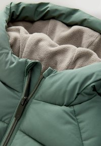 Grüne gepolsterte Jacke mit einem Reißverschluss, ausgestattet mit einem weichen, beigen Fleece-Innenfutter. Das Material ist glatt und leicht glänzend.