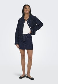 Temno modra jeans jakna s okroglim izrezom, zapiranjem na gumbe in kontrastno šivanjem, v kombinaciji z usklajenim jeans krilom. Ravne črne čevlje.