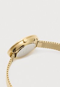 Złoty zegarek na wristwatch z siateczkowym paskiem, boczną koroną oznaczoną "ck" oraz stalowym tyłem, przedstawiony z ukośnego zbliżenia.