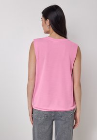 Top senza maniche di colore rosa con una texture morbida e colletto rotondo. L'orlo è leggermente curvato. Indossato con jeans grigi a vita alta.