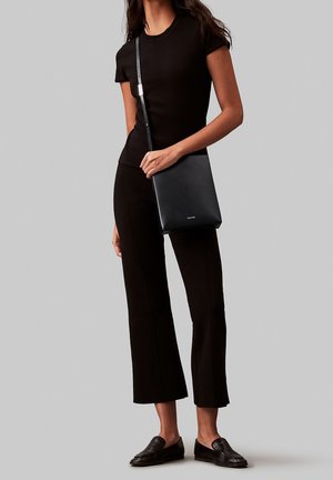 Femme en haut noir à manches courtes et pantalon portant des mocassins noirs, tenant un sac bandoulière noir sur un fond gris.