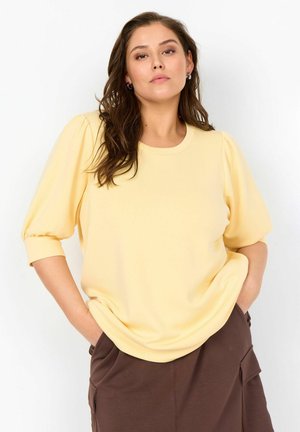 Frau mit braunen Haaren, die ein gelbes Oberteil mit Puffärmeln und braune Hosen trägt, steht mit den Händen in den Taschen vor einem einfarbigen Hintergrund.