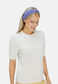 Fascia viola attorcigliata realizzata in morbido tessuto, abbinata a un maglione bianco corto con maniche a sbuffo. Design semplice, texture liscia e colori neutri.