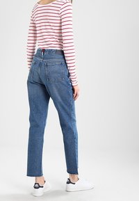 Kvinna som bär högmidjade blå jeans, vita sneakers och en vit långärmad tröja med röda horisontella ränder, stående mot en enkel bakgrund.