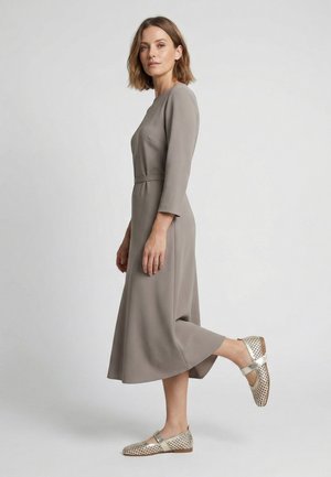 Kvinde stående på et ben iført en taupe langærmet midi-kjole og metallic vævede Mary Jane-sko mod en ensfarvet baggrund.