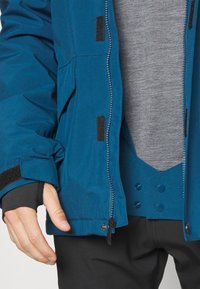 Mėlyna vandeniui atspari striukė su užtrauktuku ir Velcro užsegimais. Pasižymi lengvu dizainu, tekstūruotu audiniu ir rankų atidaromomis vietomis su thumb hole'ais.