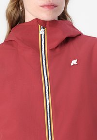 Jachetă roșie cu glugă, fermoar frontal cu dungi galbene, albastre și albe. Patch cu logo K-Way pe piept. Textură de material neted.