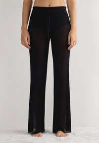 Zwarte, ribbelachtige broek met een trekkoord in de taille, met een semi-doorzichtige ontwerp en een uitlopende pijpvorm, liggend op een zachte ondergrond.