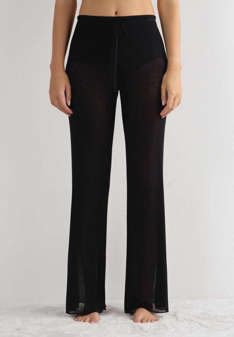 Zwarte, ribbelachtige broek met een trekkoord in de taille, met een semi-doorzichtige ontwerp en een uitlopende pijpvorm, liggend op een zachte ondergrond.