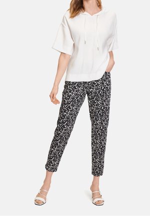 Kobieta w białej krótkiej bluzie z kapturem, czarno-białych wzorzystych wąskich spodniach i białych butach z odkrytym palcem, stoi z jedną ręką w kieszeni.