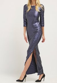 Kvinna i en lång, mörkblå glittrig klänning med trekvartslång ärm och en sprund framtill, tillsammans med svarta högklackade skor.