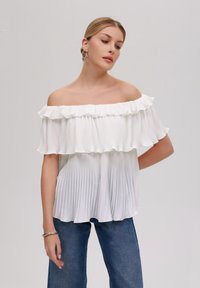 Weißes Off-Shoulder-Oberteil mit gerüschtem Ausschnitt, plissierter Textur und lockerem Schnitt, kombiniert mit blauen weit geschnittenen Jeans.