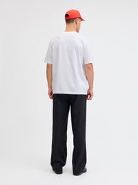 Mann trägt ein weißes T-Shirt, schwarze Nadelstreifenhose, schwarze Schuhe und eine rote Baseballkappe, steht mit dem Rücken zur Kamera vor einem weißen Hintergrund.