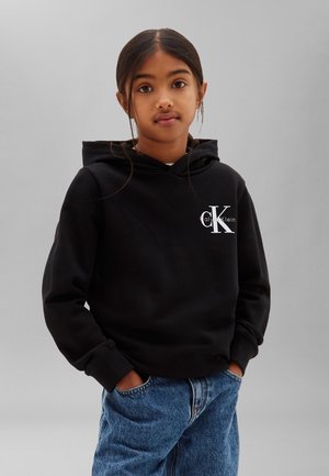 Junges Mädchen mit dunklen Haaren in einem schwarzen Calvin Klein Hoodie und blauen Jeans, steht mit den Händen in den Taschen vor einem einfarbigen grauen Hintergrund.