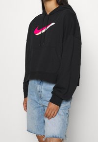 Černá zkrácená mikina s kapucí a růžovým logem Nike, kapucí na stažení a přední kapsou ve stylu klokánka, doplněná světlými džínovými šortkami.