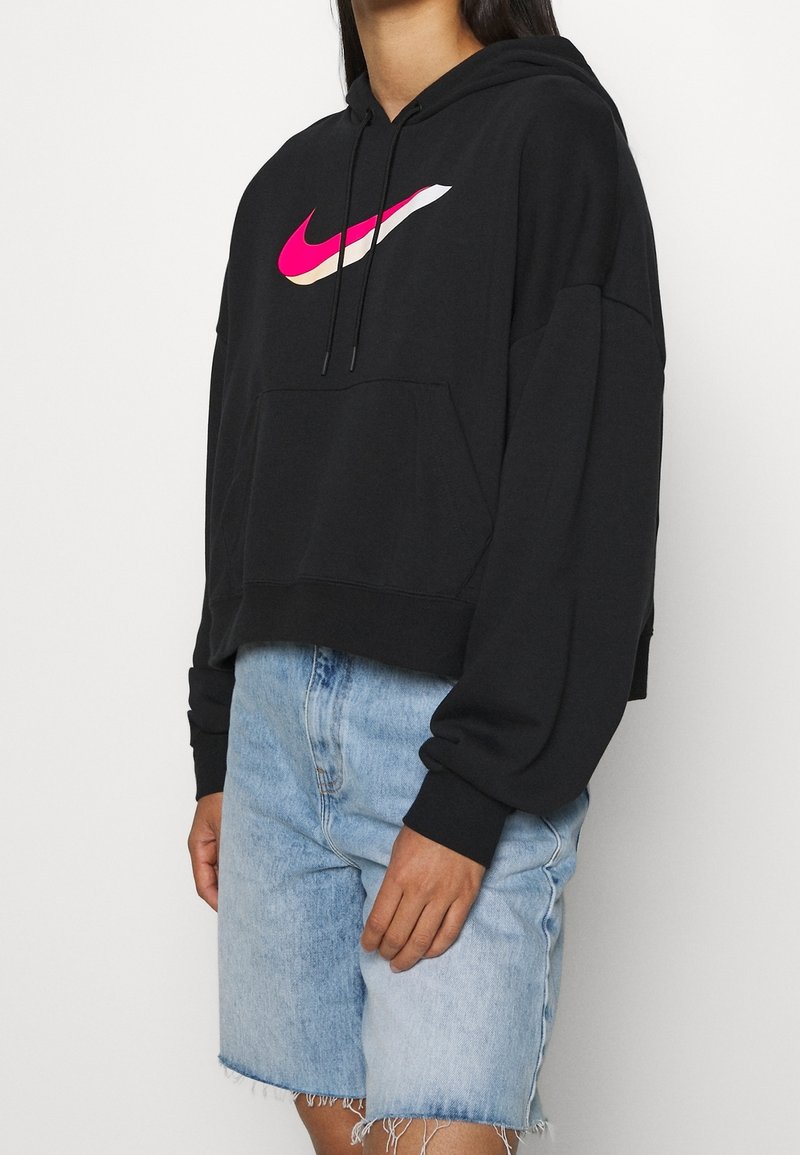 Černá zkrácená mikina s kapucí a růžovým logem Nike, kapucí na stažení a přední kapsou ve stylu klokánka, doplněná světlými džínovými šortkami.