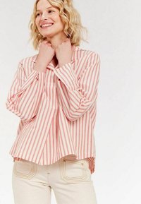 Chemise à manches longues rayée rouge et blanche avec un fit décontracté, col, et poignets boutonnés. Portée avec un pantalon de couleur claire.