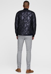 Podszyta czarna kurtka o błyszczącej fakturze, z prążkowanym kołnierzem i mankietami, noszona na granatowej bluzce i jasnoszarych spodniach, w połączeniu z brązowymi butami.