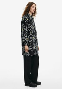 Long-sleeve, black tunic with a floral pattern in silver and yellow, paired with loose black pants and textured black shoes.