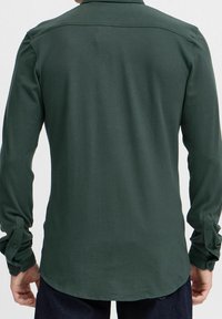 Camisa de manga larga en tela verde oscura con una textura suave, que incluye canesú en la parte trasera y puños con botones. El dobladillo es ligeramente redondeado.