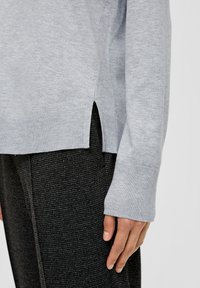 Grauer Strickpullover mit geripptem Halsausschnitt und Bündchen, seitlichen Schlitzen und darunter sichtbarem strukturiertem schwarzen Stoff.