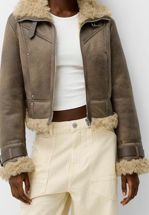 Brun shearlingjakke med pelskrave og manchetter båret over en hvid cropped top sammen med højttaljede cremefarvede bukser med forlommer.
