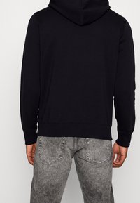 Felpa nera con cappuccio, orlo e polsini a costine, realizzata in tessuto liscio. Abbinata a jeans grigi consumati con tasche posteriori.