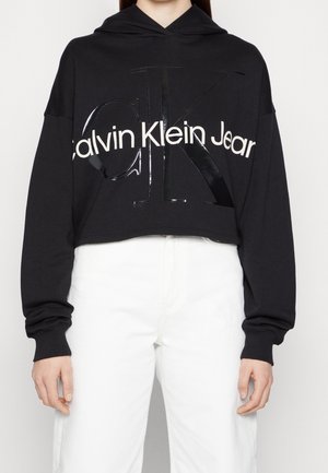 Czarna krótka bluza z kapturem z logo "Calvin Klein Jeans", noszona z wysokim stanem białych spodni, pokazująca tułów i ramiona w swobodnej pozie.