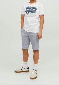 Valkoinen t-paita, jossa on sininen graafinen teksti, yhdistettynä vaaleanharmaisiin sortseihin ja valkoisiin lenkkikenkiin. Ominaisuuksiin kuuluu crew-neck-kaarroke ja rento istuvuus.