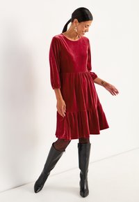 Ein rotes Samtkleid mit dreiviertel Ärmeln, rüschigem Saum und lockerer Passform, kombiniert mit knielangen schwarzen Stiefeln und durchsichtigen schwarzen Strumpfhosen.
