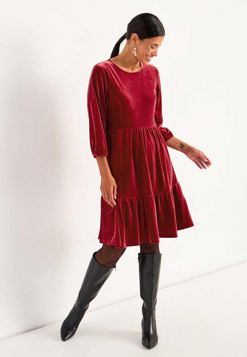 Ein rotes Samtkleid mit dreiviertel Ärmeln, rüschigem Saum und lockerer Passform, kombiniert mit knielangen schwarzen Stiefeln und durchsichtigen schwarzen Strumpfhosen.