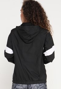 Czarna kurtka z kapturem z białymi akcentami po bokach, wykonana z lekkiego materiału, z elastycznymi mankietami i ściągaczem w talii, widziana od tyłu.