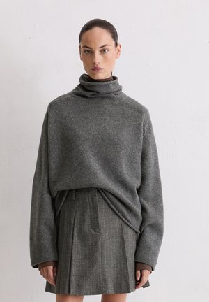 Grau gestrickter Pullover mit hohem Kragen und weiten Ärmeln, kombiniert mit einem grauen Faltenrock mit vertikalen Streifen und Taschen.