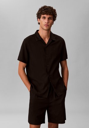 Mann mit lockigem Haar, der ein schwarzes kurzärmeliges Hemd mit Knöpfen und passende schwarze Shorts trägt, mit den Händen in den Taschen stehend.