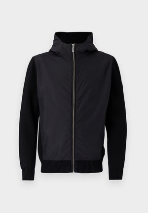 Black jacket featuring a zip front, a high collar, and mixed materials: smooth upper and ribbed knit sleeves and hem.