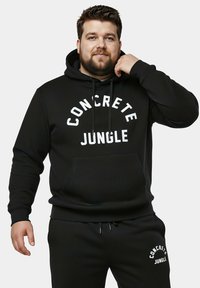Sudadera negra con un bolsillo frontal y texto en blanco "CONCRETE JUNGLE". El material parece texturizado, combinado con pantalones de chándal a juego que muestran un logo.