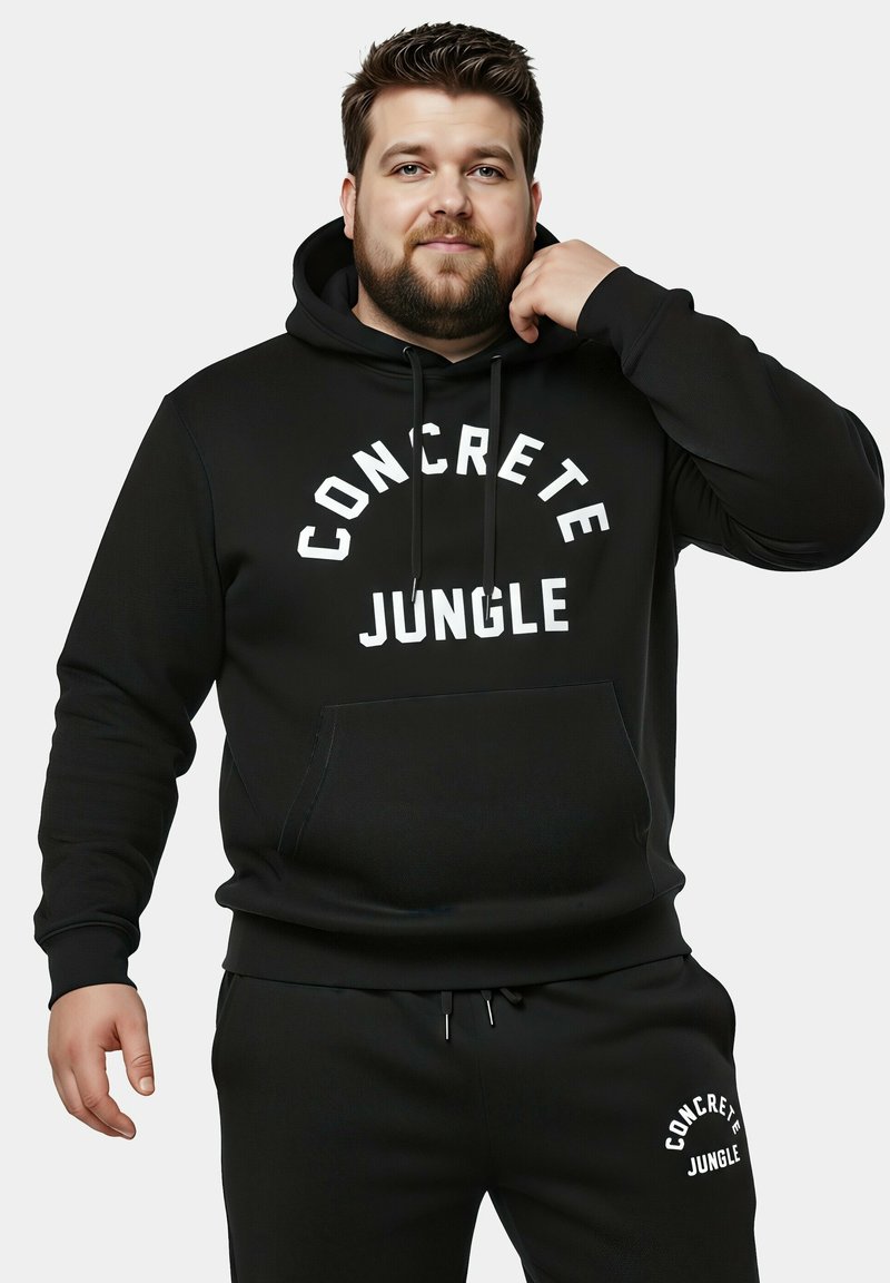 Sudadera negra con un bolsillo frontal y texto en blanco "CONCRETE JUNGLE". El material parece texturizado, combinado con pantalones de chándal a juego que muestran un logo.