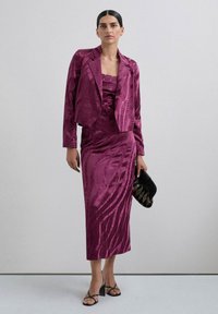 Traje de satén fucsia con chaqueta corta y vestido ajustado con patrones ondulados. Completado con sandalias de tiras y un bolso de mano negro.